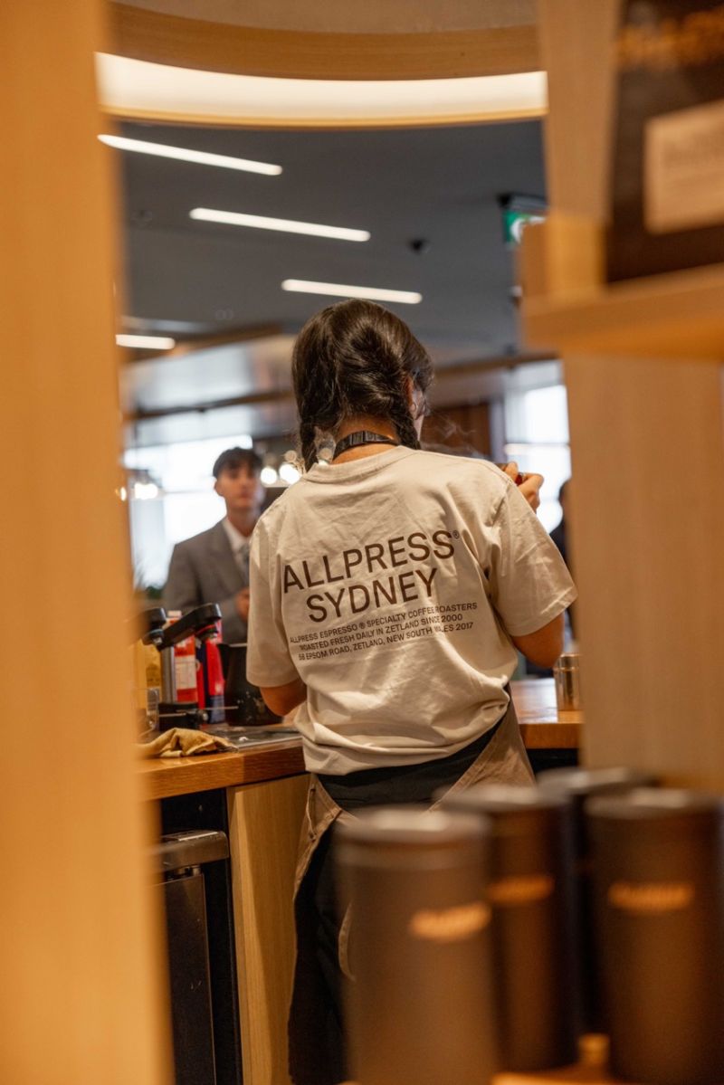 A person standing at a cafe counter seen from behind, wearing a white graphic T-shirt with 'ALLPRESS SYDNEY' printed in black across the back.